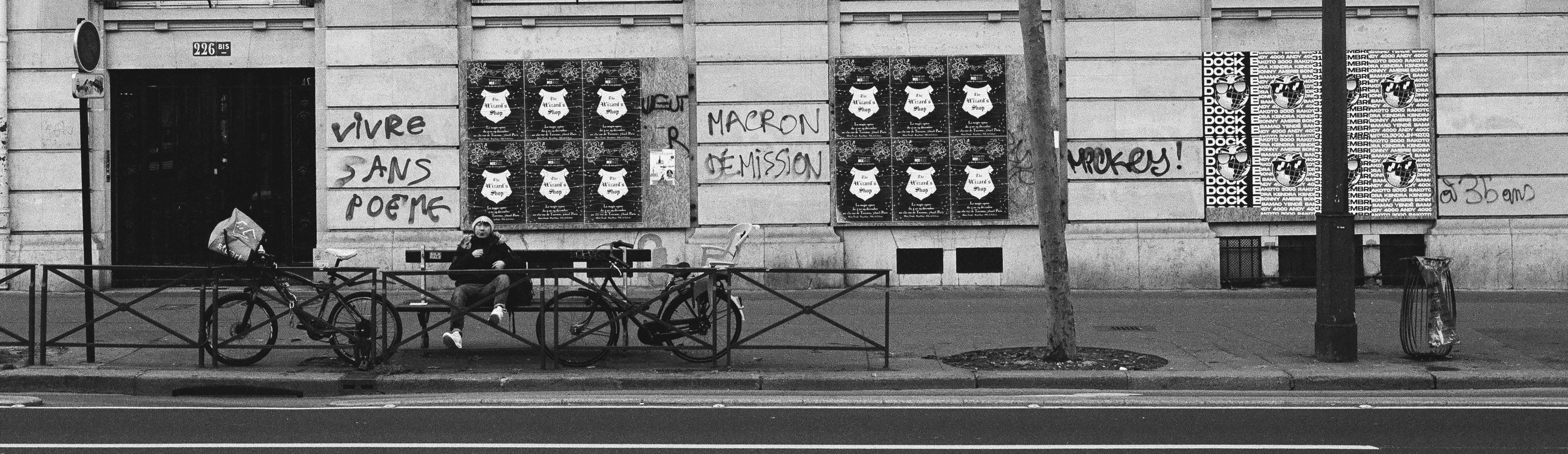black and white photo of french street, graffiti on walls, man sitting on bench near two bicycles